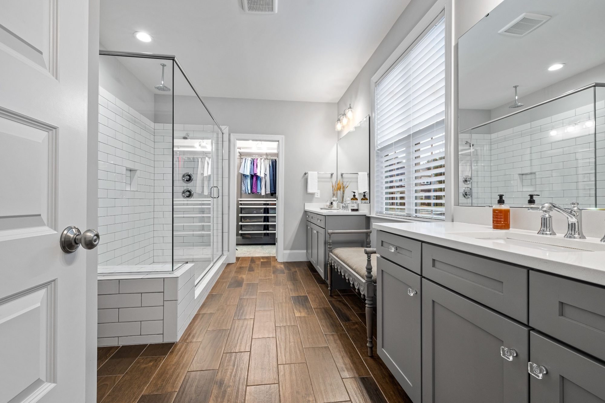 Stock image of modern bathroom with gray vanities, glass shower, white tile, and wood-look flooring -optimized