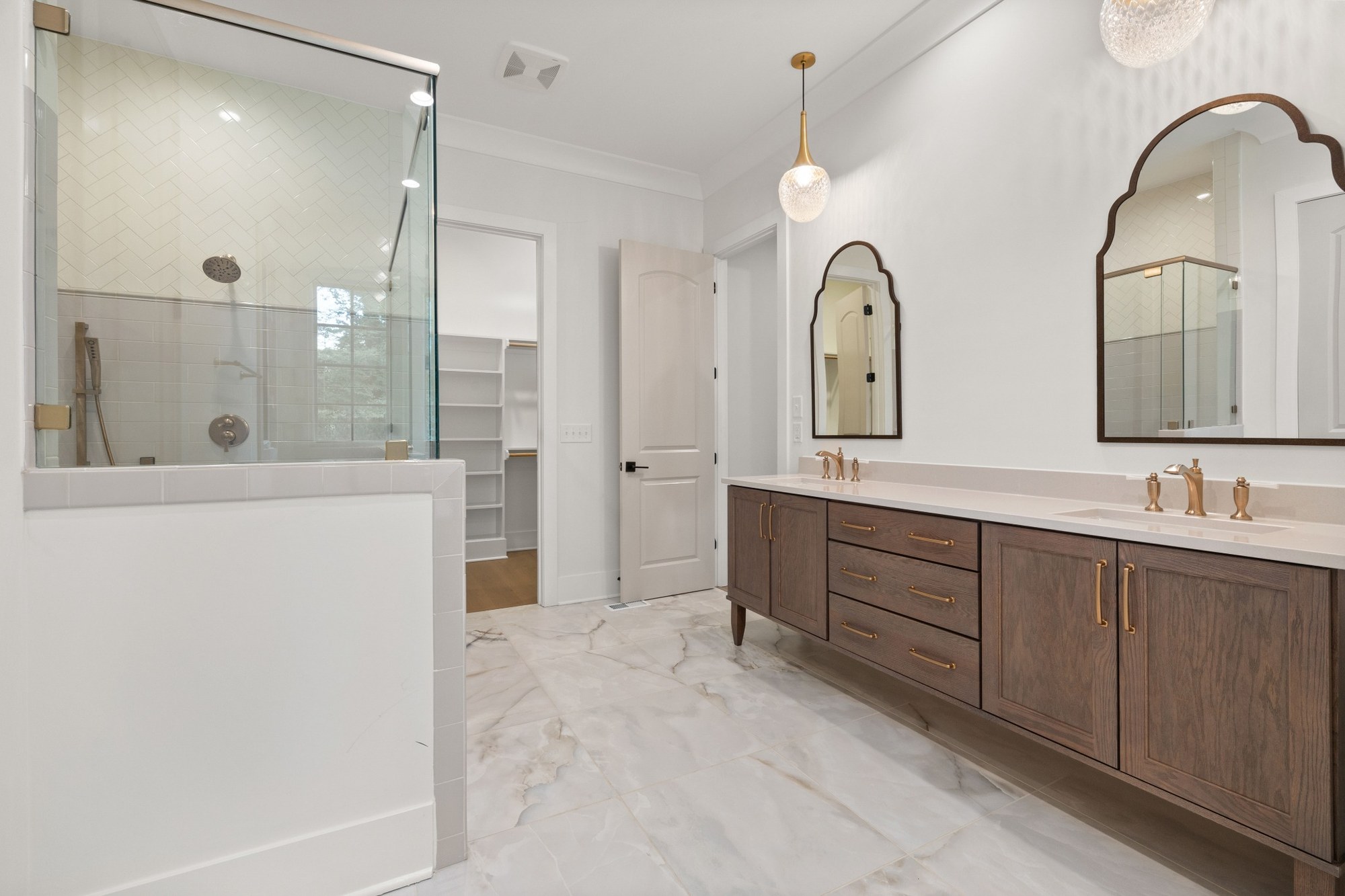 Stock image of modern bathroom with wood double vanity, gold fixtures, glass shower, and marble tile -optimized